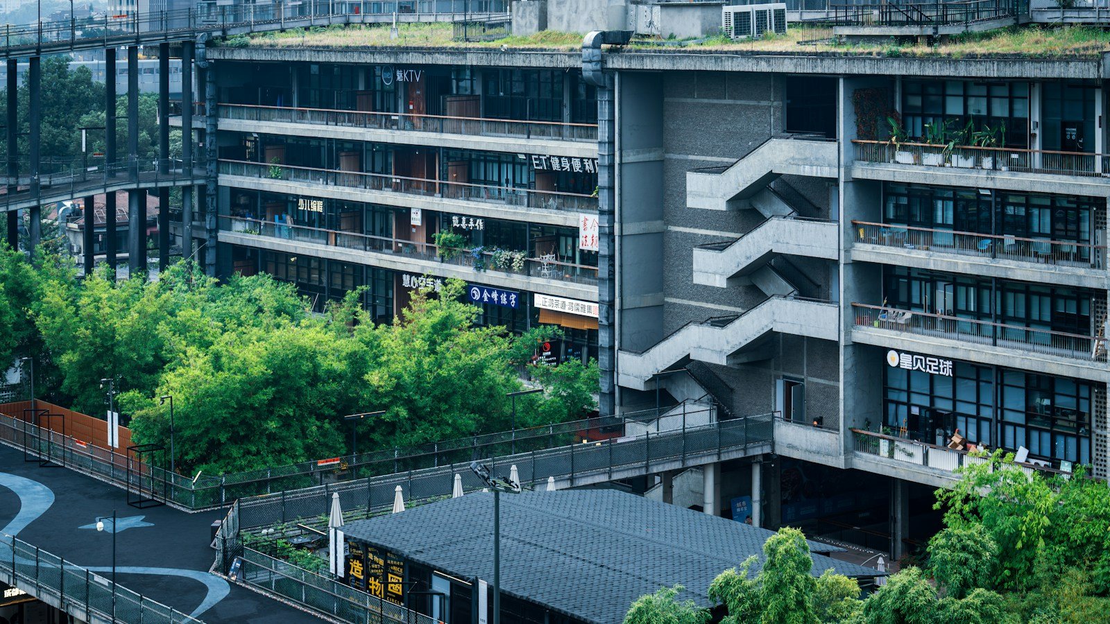 Edificio moderno a più piani con tetto verde e terrazze piantumate, circondato da alberi e bambù lungo una strada
