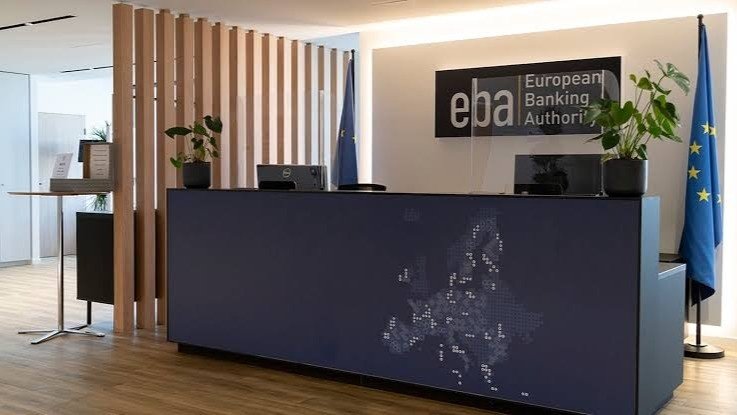 Bancone della reception dell'Autorità bancaria europea con l'insegna “eba European Banking Authority” alla parete, due bandiere dell'Unione europea e una mappa d'Europa a puntini sul fronte del bancone