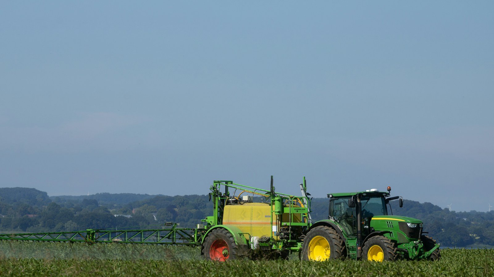 Trattore verde con irroratrice gialla e barra aperta che attraversa un campo coltivato, colline boscose sullo sfondo